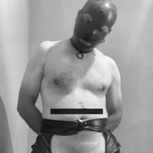 Hombre con el torso desnudo posando en blanco y negro dentro de un espacio interior sencillo, con una máscara negra de cuero tipo bondage que cubre toda la cabeza y deja aberturas para los ojos y la boca. Lleva un collar ajustado con una anilla metálica en el cuello, las manos colocadas detrás de la espalda y una prenda de cuero oscuro ceñida a la cintura, con hebillas y cordones visibles. Una barra negra censura la zona baja del abdomen y el pubis, reforzando el carácter fetichista y erótico de la escena. La iluminación suave resalta el pecho, los hombros y la textura brillante del cuero, creando una atmósfera íntima, austera y dominada por estética BDSM.