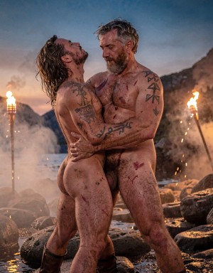 Amleth y Aurvandil.  Versión cinematográfica explícita de la imagen: Dos guerreros vikingos adultos de cuerpo entero se aferran en una pose ritual intensa sobre la orilla rocosa de un fiordo nórdico al anochecer, con el guerrero de 29 años delante arqueando la cabeza hacia atrás y el rey guerrero de 48 años sujetándolo por la cintura desde atrás. La niebla baja se arrastra entre piedras húmedas, antorchas reales lanzan chispas y humo ondulante, y la piel muestra poros, barro agrietado, sangre seca, cicatrices elevadas, tatuajes rúnicos oscuros, vello corporal, cabello enmarañado y barbas sucias. El contraluz crepuscular profundo dibuja un degradado de azul cobalto a naranja quemado, mientras una iluminación cinematográfica de tres puntos añade clave alta controlada y relleno lateral cálido sobre los torsos. La composición vertical 9:16 usa lente anamórfica de 40mm, grano analógico tipo Kodak Vision3 500T, contraste HDR con sombras profundas y altas luces contenidas, en un realismo brutal de saga nórdica.