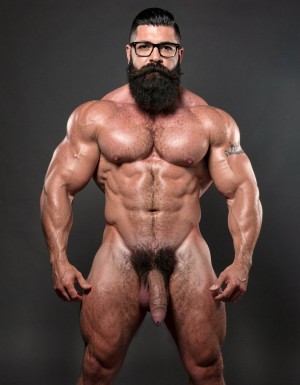 A highly detailed full-body nude photograph of a 40-year-old dominant muscle bear named Hipsterbarba, standing confidently in a well-lit studio with dramatic lighting that accentuates every muscle. He is 180cm tall, 80kg with 8% body fat, extremely muscular and symmetrical. Massive rounded hairy pecs with clear separation and dark pointed nipples, shredded six-pack abs with deep cuts and visible obliques forming a strong V-taper, wide shoulders, vascular arms, huge quads and defined legs. Very dense black masculine body hair covering his chest, running down in a thick treasure trail to a very bushy dark pubic area. He has a perfectly groomed long Bandholz beard that reaches mid-chest, thick mustache with curled upward spiral ends, modern high fade undercut hairstyle, black hipster glasses, and a small tattoo on his right bicep that reads 