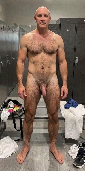 A bald man poses standing and completely naked in a locker room, facing the camera with a neutral expression and straight posture, arms relaxed at his sides and legs slightly apart. He has a muscular and athletic body, with chest, abdomen, pubis, and legs covered in abundant body hair; his penis is visible in the center of the image. The setting is a locker room with gray metal lockers on both sides, a bench, tiled floor, and disordered sportswear, including white towels, colored garments, and some sneakers next to the bench, which reinforces a context of gym, shower, or changing clothes.