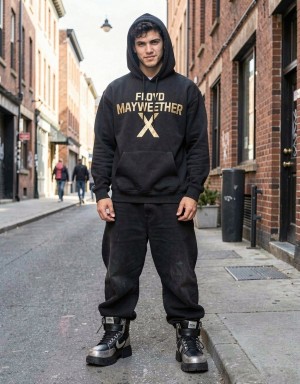 A young man standing in the middle of a narrow urban street poses facing forward with a calm attitude, wearing a black hoodie with the hood up and the text “Floyd Mayweather” next to a large golden “X” on the chest. He wears loose black sporty-style pants and tall black boots with gray details, giving a casual streetwear look. The background shows brick buildings and old façades on both sides, a stone sidewalk, dark doors and windows, as well as several people walking blurred in the background, reinforcing the atmosphere of a central neighborhood, urban fashion, and outdoor portrait photography.
