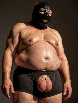 Hombre corpulento y desnudo de torso, con barriga prominente, pecho ancho y brazos robustos, posando de pie contra un fondo oscuro y sobrio. Lleva una máscara negra de cuero que cubre casi toda la cabeza y deja visibles los ojos, aportando una estética fetichista y BDSM. Viste unos calzoncillos bóxer negros ajustados con una abertura frontal por la que sobresalen claramente el pene y los testículos, centrando la atención en la zona genital. La iluminación lateral resalta la piel, el volumen del abdomen, los pliegues corporales y la textura brillante de la máscara, creando una escena explícita, directa y de carácter erótico.