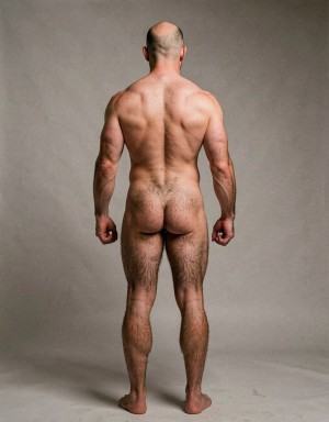 Mike Morris, a 32-year-old extremely lean Caucasian male at only 8% body fat, stands completely naked in a strict military attention pose seen from directly behind in a full posterior view. His back is perfectly straight, shoulders pulled back, arms rigid at his sides with fists clenched, legs straight and together, head held high. The shot captures him from the heels of his feet all the way up to the top of his bald head. His advanced Norwood III-IV baldness is clearly visible from behind, with a smooth tanned scalp on the crown and short dense dark brown hair only on the sides and lower back of the head. His back is highly defined and vascular: wide lats tapering dramatically to a very narrow, tight waist, thick trapezius muscles, and dense dark brown body hair lightly covering the shoulders and becoming much thicker and denser across the lower back and lumbar area, especially between the lats. His ass is tight, muscular and round with almost no fat, the glutes clearly separated. Thick dark hair continues down onto his powerful diamond-shaped calves and the backs of his slightly hairy feet. Dramatic directional studio lighting from above and slightly to the side creates strong highlights on his dry, shredded skin and deep shadows that accentuate every muscle fiber, the texture of his dense back hair, and the extreme vascularity and definition of his 8% body fat physique. Photorealistic, high-contrast, masculine 1970s Colt Studio aesthetic, grainy film texture, commanding and powerful presence. Las piernas están un poco abiertas pies un poco más separdos