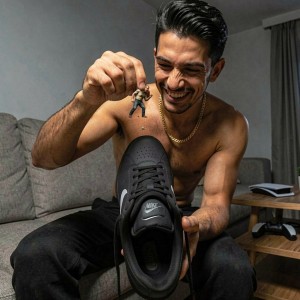 A smiling shirtless man appears sitting on a gray living room sofa, wearing black pants and a gold chain around his neck, playing with perspective as he holds a small human figure between his fingers over a black Nike sneaker placed in the foreground. The scene has a fun and creative tone, as if the tiny character were about to fall onto the sports shoe, whose laces, logo, and perforated texture are shown in great detail. In the background there is a wooden side table, a notebook or pad, a video game controller, and gray curtains, creating a casual and modern domestic atmosphere.