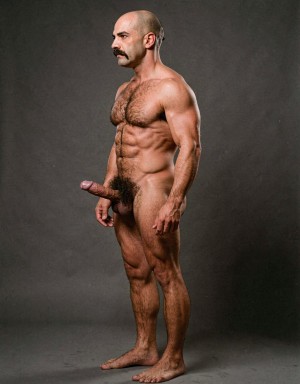 Mike Morris, a 32-year-old extremely lean Caucasian male with only 8% body fat, stands completely naked in a strict military attention pose captured in a full-body exact side profile at 90 degrees from his left side. The shot runs from the tips of his size 44 slightly hairy feet all the way up to the top of his bald head, showing his entire body without any cropping. His physique is dry, highly vascular and powerfully defined: massive square pectorals seen in perfect side silhouette, dense dark brown chest hair forming a thick T-pattern, sharply carved six-pack abs in clear profile, narrow waist, wide lats, powerful thighs and diamond-shaped calves. Dramatic directional studio lighting comes from high and slightly in front of him, creating strong rim lighting that traces the sharp outline of his entire body, the texture of his dense dark body hair on chest, lower back, ass and legs, and deep shadows that accentuate every muscle separation, vein and the extreme dryness of his 8% body fat physique. His advanced Norwood III-IV bald scalp shines under the light, with short dense dark brown hair on the sides and back of his head. His square angular face is shown in pure profile: strong jawline, prominent chin with slight central dimple, thick dark handlebar mustache, heavy brow and intense deep-set brown eye looking straight forward with a serious, commanding expression. His penis is fully visible in perfect side view, completely natural and realistic: thick, 24 cm long when fully erect, with a natural slight upward curve, prominent dorsal veins, long foreskin partially covering the pink-red glans, and realistic lighter skin tone. Below it hang his large, low-hanging testicles with wrinkled darker skin and dense dark brown hair. The extremely thick, curly dark brown pubic bush climbs almost to his navel and surrounds the base of his cock and balls generously. Every anatomical detail from his veined hands and forearms to his muscular ass, hamstrings, diamond calves and hairy toes is perfectly proportioned and free of any deformation. Photorealistic, high-contrast, masculine 1970s Colt Studio aesthetic with subtle grainy film texture.