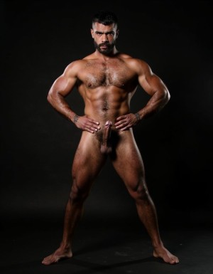 Un hombre musculoso y dominante de 35 años, completamente desnudo, cuerpo atlético y marcado, piel morena, brazos tatuados, pecho ancho y velludo, abdominales definidos, piernas musculosas, polla grande y gruesa erecta, mirada intensa y arrogante directa a cámara, expresión dominante y segura, de pie con las manos en las caderas en posición de poder, iluminación dramática que resalta sus músculos, fondo oscuro, fotografía hiperrealista, calidad 8K, estilo fotográfico profesional masculino.