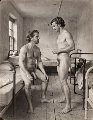 Two naked men pose in an austere, old-looking room, probably a shared bedroom, hospital, or barracks, with metal beds on both sides, thin mattresses, folded blankets, a wooden floor, and a high window illuminating the black-and-white scene. The man on the left is sitting on the edge of a bed, with his legs apart, his hands resting near his knees, and his penis visible, looking at the other with an attentive expression. The man on the right remains standing, also naked, with his penis visible, his torso slightly turned and one hand raised as if conversing or explaining something. The photograph has a documentary and vintage air, with worn walls, natural light, and an intimate but sober composition.