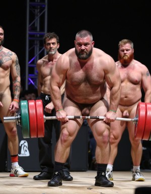 Varios hombres extremadamente musculosos y barbudos en una competición de halterofilia. uno de ellos, desnudo, levanta una enorme barra con pesas de trescientos kilos