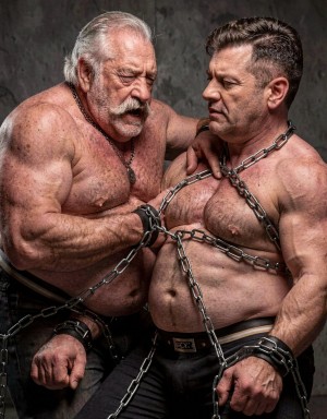 Two burly, muscular men appear shirtless in a rough and dominant-looking scene, with metal chains crossed over their chests, necks, and arms, as well as black leather cuffs on their wrists. The man on the left, older, with white hair combed back, a thick mustache, and a hairy torso, tilts his face with an intense expression while resting one hand on the other man’s shoulder. The man on the right, with short dark hair and a strong jawline, looks sideways with a serious, defiant expression, showing a strong abdomen and hairy chest. The dark concrete background, dramatic lighting, and bondage accessories create a masculine, tense, fetishistic atmosphere.