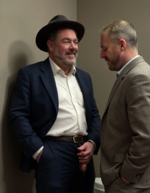 Two men in suits stand talking in an interior corner with beige walls, in a discreet and somewhat intimate atmosphere, possibly an office, hallway, or private room. The man on the left wears a navy blue suit, an open white shirt without a tie, a dark belt, and a short-brimmed black hat; he has a graying beard, a smiling and relaxed expression, one hand in his pocket and the other resting on his lower abdomen. The man on the right wears a light gray suit with a white shirt, maintains a close posture with one hand in his pocket, and looks down while smiling. The soft lighting creates shadows on the wall and reinforces the informal tone of the conversation between them.