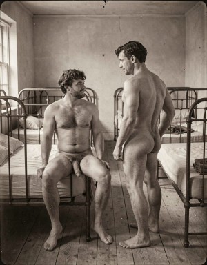 Dos hombres desnudos aparecen en una habitación austera de aspecto antiguo, con camas metálicas alineadas a ambos lados, colchones claros, suelo de madera y una ventana que deja entrar luz lateral. Uno está sentado en el borde de una cama, con barba, pelo rizado, torso velludo y las piernas abiertas, mostrando el pene en reposo mientras mira al otro con gesto serio. El segundo hombre permanece de pie de espaldas y ligeramente girado, con el cuerpo musculado, las nalgas visibles y la cabeza inclinada hacia su compañero. La escena, en blanco y negro con tono sepia, transmite una atmósfera íntima, silenciosa y casi documental, propia de un dormitorio colectivo o barracón.
