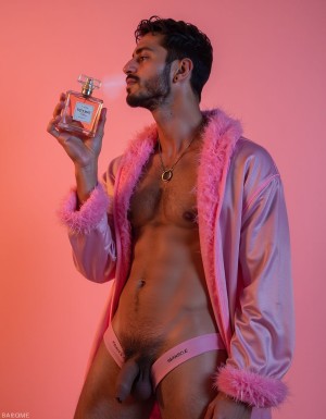 Man posing with a sensual attitude against a pink background lit in warm tones, dressed in an open pink satin robe with fuchsia faux-fur trim on the collar and cuffs, exposing his torso, defined abdomen, and body hair. He wears a pink Barcode jockstrap or underwear that leaves the penis and scrotum visible at the front, along with a gold chain with pendant, earring, and trimmed beard. He holds a square perfume bottle with pink liquid and a metallic cap near his face, looking to the side with an elegant and provocative expression, in a fashion, fragrance, queer glamour, and studio photography aesthetic.